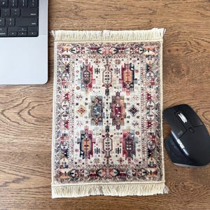 May include: A rectangular mousepad with a traditional rug design. The pad features a cream-colored background with intricate patterns in red, blue, orange, and black. The edges are finished with a fringe.