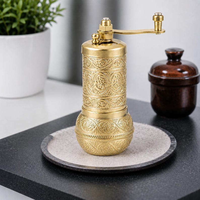 Puede incluir: Un molinillo de especias cil&iacute;ndrico de lat&oacute;n con un intrincado patr&oacute;n floral en relieve en todo el cuerpo. El molinillo incluye una manivela manual montada en la parte superior con un peque&ntilde;o pomo, una base redondeada y un acabado dorado met&aacute;lico.