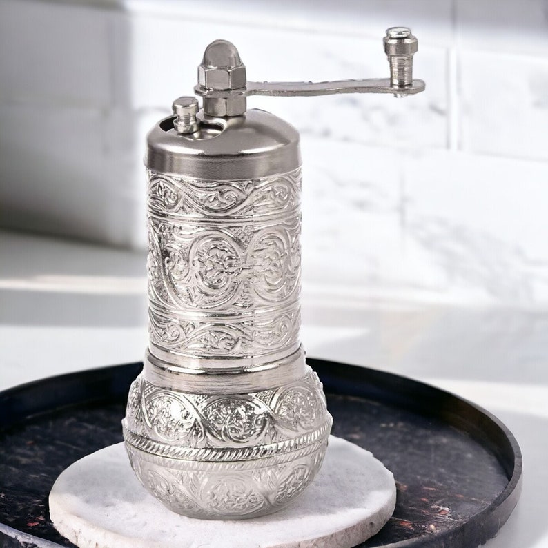 Puede incluir: Un molinillo de especias de metal plateado con un intrincado patr&oacute;n floral en relieve en su cuerpo cil&iacute;ndrico y base redondeada. La parte superior incluye una manivela manual y un peque&ntilde;o pomo para ajustar el molido, con acabado met&aacute;lico.