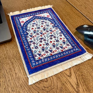 May include: A small prayer rug with a detailed floral design in red, blue, and white. The rug has a blue border and fringe along the edges. The design features a central arch shape with floral and bird motifs.
