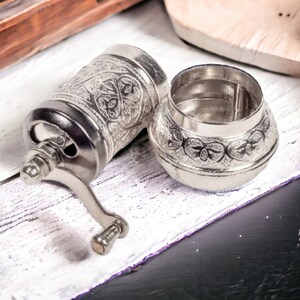 Puede incluir: Un molinillo de especias de metal color plata con un intrincado patr&oacute;n floral en relieve en el cuerpo cil&iacute;ndrico y la tapa desmontable. El molinillo cuenta con una manivela manual con un peque&ntilde;o pomo y una amplia abertura superior.