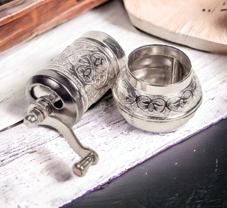 Puede incluir: Un molinillo de especias de metal color plata con un intrincado patr&oacute;n floral en relieve en el cuerpo cil&iacute;ndrico y la tapa desmontable. El molinillo cuenta con una manivela manual con un peque&ntilde;o pomo y una amplia abertura superior.