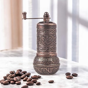 Puede incluir: Un molinillo de caf&eacute; cil&iacute;ndrico de metal en tono cobre con un intrincado patr&oacute;n floral en relieve en todo el cuerpo. El molinillo incluye una manivela manual montada en la parte superior y est&aacute; rodeado de granos de caf&eacute; tostados.