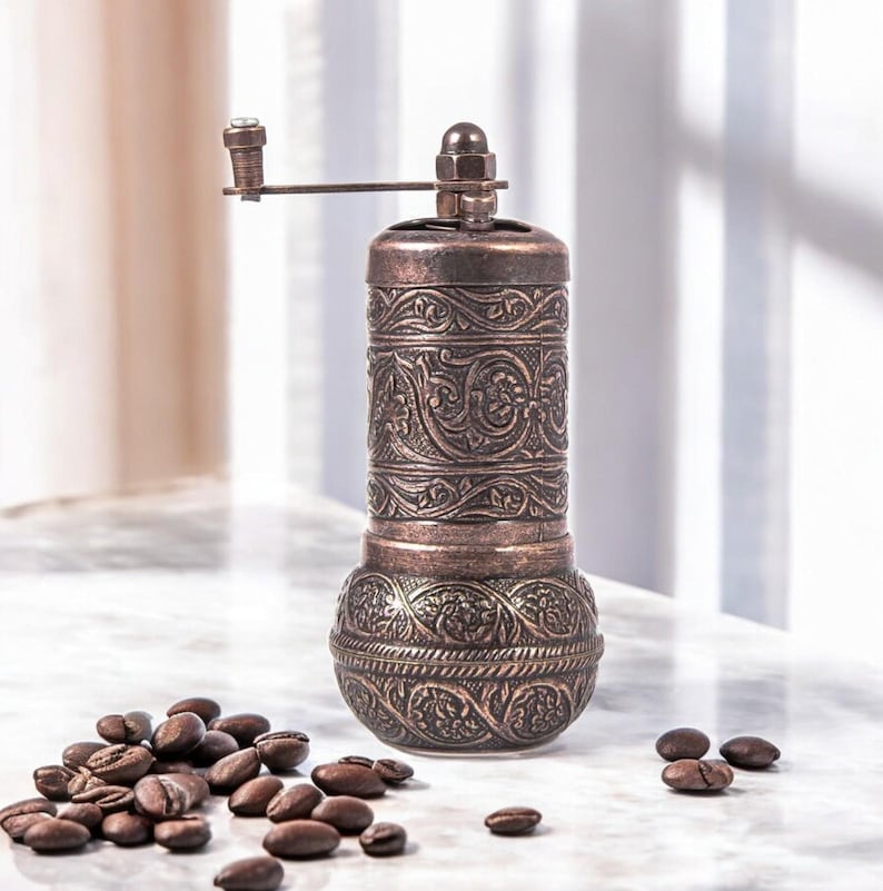 Puede incluir: Un molinillo de caf&eacute; cil&iacute;ndrico de metal en tono cobre con un intrincado patr&oacute;n floral en relieve en todo el cuerpo. El molinillo incluye una manivela manual montada en la parte superior y est&aacute; rodeado de granos de caf&eacute; tostados.