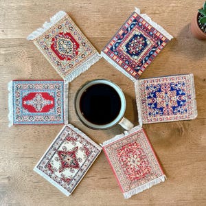 Op de afbeelding: Zes miniatuur tapijt onderzetters met gedetailleerde patronen in rood, blauw en crème, rondom een koffiekopje. Elke onderzetter heeft een gefranjerde rand, die traditionele tapijten nabootst. Geplaatst op een houten oppervlak.
