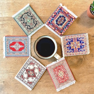 May include: A set of six rectangular drink coasters featuring intricate woven rug patterns in various colours including red, blue, green, and cream, each finished with white fringe on the short ends, arranged around a white ceramic mug of black coffee.