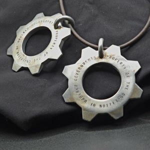 Puede incluir: Dos colgantes plateados en forma de engranaje con la inscripción "PROPERTY OF THE COALITION OF UNDERGROUND GOVERNMENTS" grabada en el círculo interior. Uno está en un collar de cordón marrón, sobre un fondo oscuro.