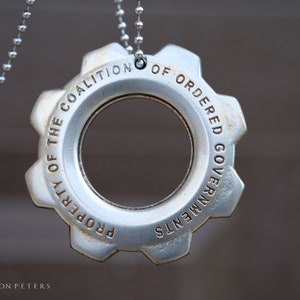 Puede incluir: Un colgante de engranaje plateado con la inscripción "Property of the Coalition of Ordered Governments" colgando de una cadena plateada.