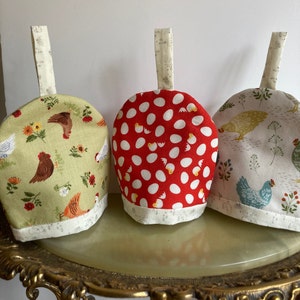 Peut inclure: Trois housses de thé en tissu avec des motifs différents. La première est verte avec un motif de poulet blanc et floral. La deuxième est rouge avec des pois blancs et un motif d'œuf blanc. La troisième est beige avec un motif de poulet bleu clair et floral.