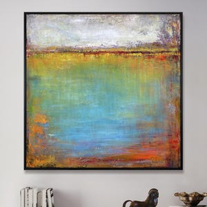 Op de afbeelding: Abstract landschapsschilderij in een zwarte lijst. Het kunstwerk toont een mix van kleuren, waaronder blauw, groen, geel en oranje, waardoor een serene en gestructureerde uitstraling ontstaat. Het schilderij wordt tentoongesteld boven een moderne witte en houten kast.