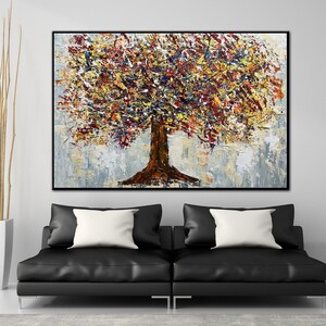 May include: An abstract tree painting with a black frame. The tree trunk is brown, and the leaves are a mix of yellow, red, and orange. The background is a light gray and white. The painting is displayed above two black leather sofas with white pillows.