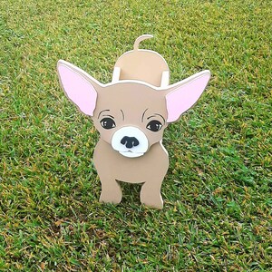 May include: A brown wooden chihuahua figurine with pink ears. The figurine is standing on a green grassy surface.