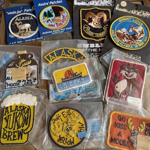 May include: A collection of twelve embroidered fabric patches featuring various Alaska and Yukon themes, including depictions of moose, sheep, Bigfoot, and scenic landscapes, with some patches still attached to their original cardboard backing.