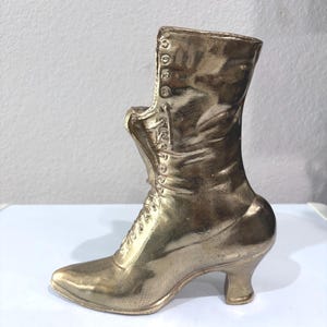 May include: A gold-colored, decorative boot with a pointed toe and a small heel. The boot features a lace-up design and button details along the side. The boot is a vintage-inspired piece, likely a decorative item.
