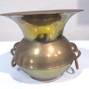 May include: A vintage brass vase with a flared rim and two decorative handles. The vase has a bulbous body and a warm, golden hue. The rim is wide and slightly angled, and the handles are small and circular.