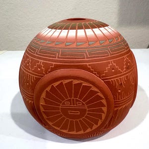 May include: A terracotta-colored, spherical ceramic vase with intricate geometric patterns and a sun-like design. The vase features a small opening at the top and is adorned with detailed carvings, showcasing a Southwestern or Native American artistic style.