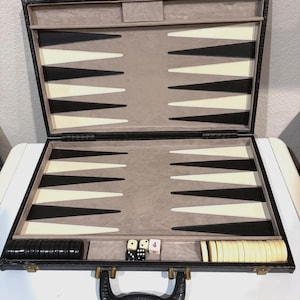 May include: An open backgammon board game set in a black carrying case with a textured exterior. The interior features a grey felt surface with alternating black and white triangular points. Includes black and white checkers, two dice, and a doubling cube.