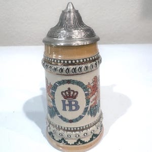 May include: A ceramic beer stein with a conical metal lid featuring an embossed floral pattern. The body is cream-colored with a central blue HB logo topped by a red crown, flanked by two orange lions within a green wreath. Decorative bands border the top and bottom.