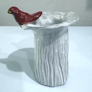 May include: A white ceramic vase with a textured, tree-bark-like surface and a wide, ruffled rim. A small, glossy red bird figurine with a yellow beak is perched on the edge of the rim, integrated into the structure of the vessel.