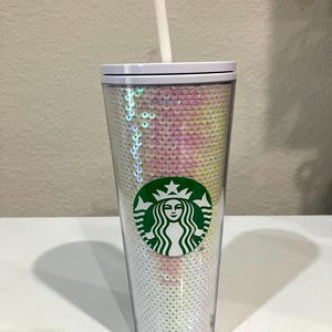 May include: A white reusable tumbler with a green Starbucks logo and a clear plastic lid. The tumbler is covered in iridescent sequins that change color in the light.
