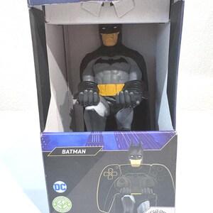 May include: A Batman figure controller and phone holder packaged in a rectangular box. The figure features a grey and black suit with a yellow utility belt. The box displays the Batman logo, the text BATMAN, DC, and THE ORIGINAL PHONE AND CONTROLLER HOLDER.