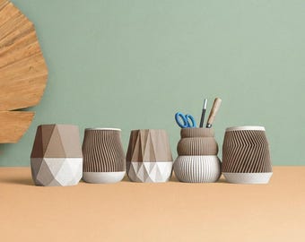 May include: Five small containers in various brown and white geometric designs arranged in a row. The containers feature textured surfaces with vertical ridges, faceted patterns, and smooth bands. One container holds blue scissors and two pens.