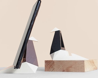 May include: Two white origami phone stands with a black smartphone in one of the stands. The stands are on a wooden base.
