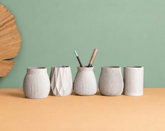 May include: Five small, light gray cylindrical containers with various textured surfaces, including vertical ribs, geometric facets, and smooth finishes, arranged in a row with two thin writing instruments placed inside the center vessel.