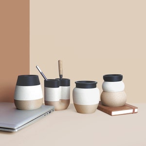 Puede incluir: Cinco tazas de cerámica con diseños en blanco y negro. Las tazas están dispuestas en una fila sobre una superficie marrón claro. Un ordenador portátil está en primer plano.