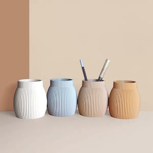 Peut inclure: Quatre pots cylindriques rainurés pour le bureau, blancs, bleu clair, beige et orange. Deux stylos sont dans le pot beige. Les pots sont sur une surface beige clair avec un fond bicolore.