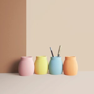 Op de afbeelding: Vier kleurrijke, geribbelde, cilindrische containers in roze, geel, blauw en oranje. De containers staan in een rij op een lichtbruin oppervlak.