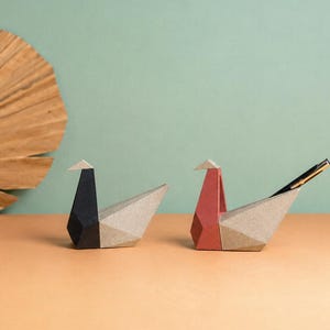 May include: Two geometric bird-shaped desk organizers made of folded material. One bird is black and light gray, while the other is red and light gray. The red and gray bird holds a black pen in its back section, showcasing a functional design.