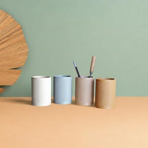 Peut inclure: Quatre porte-stylos cylindriques avec une texture nervurée verticale, disposés en rangée. Les contenants sont blancs, bleu clair, marron clair et beige. Deux instruments d'écriture sont placés dans le porte-stylo marron clair, le troisième.
