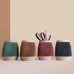 Peut inclure: Quatre porte-stylos décoratifs verts, marron, rouges et bleu marine. Chaque support présente un motif en chevrons et une base en liège. Un support contient des ciseaux, un crayon et un stylo.