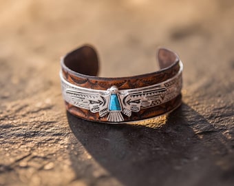 May include: A cuff bracelet featuring a brown leather band with an embossed pattern and a central silver-toned metal overlay shaped like a bird with outstretched wings, inlaid with a small, rectangular piece of bright blue turquoise stone.
