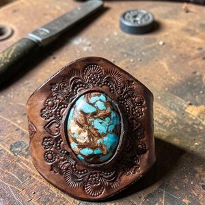 May include: A brown leather cuff bracelet featuring an oval turquoise stone with brown matrix veining. The leather surface is decorated with intricate, symmetrical floral and heart-shaped embossed patterns surrounding the central stone setting.