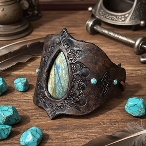 May include: A wide dark brown leather cuff bracelet featuring an embossed floral pattern and a large central teardrop-shaped labradorite gemstone with blue and yellow iridescence, accented by small round turquoise stones on the sides.
