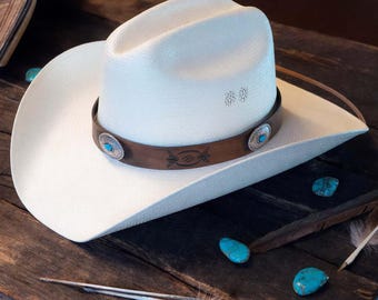 Könnte beinhalten: Ein weißer Cowboyhut mit einem braunen Lederband und türkisfarbenen Akzenten. Der Hut hat eine gewebte Textur und ist mit kleinen silbernen Verzierungen verziert. Der Hut wird auf einer rustikalen Holzoberfläche mit Türkissteinen und Federn präsentiert.