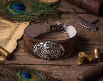 May include: Brown leather cuff bracelet with a silver decorative centerpiece. The bracelet has a buckle closure and is surrounded by peacock feathers, bullets, and a vintage map on a wooden surface.