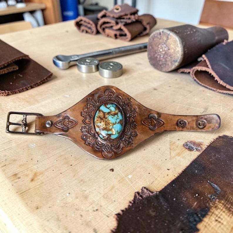 May include: A brown leather cuff bracelet featuring an oval turquoise stone set in the center. The leather is embossed with intricate floral patterns and includes a metal buckle closure with multiple adjustment holes for a secure fit.