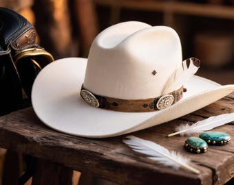 Könnte beinhalten: Ein cremefarbener Cowboyhut mit einem dekorativen braunen Lederband und silbernen Verzierungen. Eine einzelne Feder und zwei türkisfarbene Steine mit goldenen Akzenten liegen auf einer Holzoberfläche.