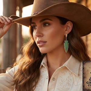 May include: A woman wearing a brown cowboy hat and a cream-colored shirt with floral embroidery. She is wearing large, green arrowhead-shaped earrings with gold accents. The image includes the Gray Wolf Collections logo.