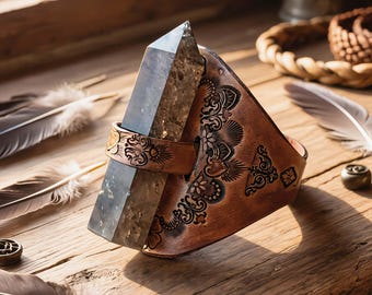 May include: A brown leather crystal holder with intricate floral and geometric patterns. A large, faceted, smoky quartz crystal is held in place by a leather strap. The holder sits on a wooden surface with feathers.