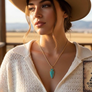 May include: A gold-colored necklace with a turquoise arrowhead pendant. The woman is wearing a cream-colored sweater and a tan hat. The Gray Wolf Collections logo is in the bottom right corner.