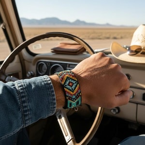 Puede incluir: Una pulsera ancha de cuentas con un patrón geométrico en tonos vibrantes de turquesa, rojo, amarillo y negro. La pulsera se lleva en una muñeca sobre un volante, con un sombrero de vaquero de paja y gafas de sol visibles en el salpicadero.