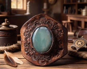 May include: A brown leather cuff bracelet with an oval, light green stone at the center. The leather is embossed with floral and scroll designs. A gold-colored sheriff's badge and a feather are on the wooden surface.
