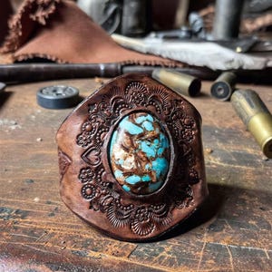 May include: A brown leather cuff bracelet featuring an oval turquoise stone with brown matrix veining at the center. The leather band is decorated with an embossed floral and heart pattern surrounding the stone, creating a textured surface.
