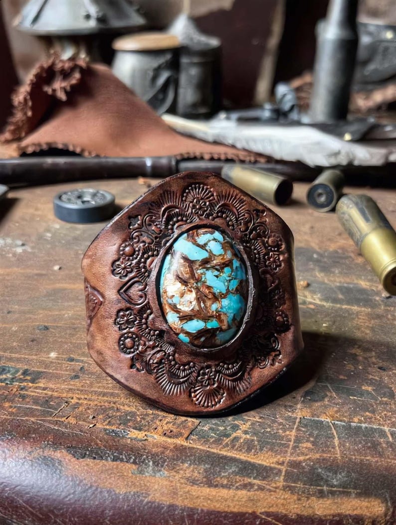 May include: A brown leather cuff bracelet featuring an oval turquoise stone with brown matrix veining at the center. The leather band is decorated with an embossed floral and heart pattern surrounding the stone, creating a textured surface.