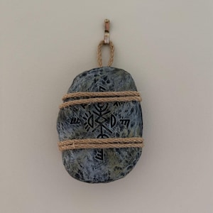 May include: A gray stone with a black geometric design is wrapped with brown twine and hung from a hook on a white wall.
