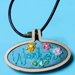May include: Oval pendant necklace with a light blue fabric background. The word "Wander" is embroidered in teal thread, surrounded by colorful embroidered flowers. The pendant is framed in wood and hangs from a black cord.